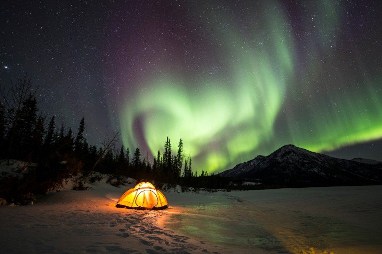 Aurora boreal verde sobre una tienda amarilla iluminada junto a un río congelado durante la noche.