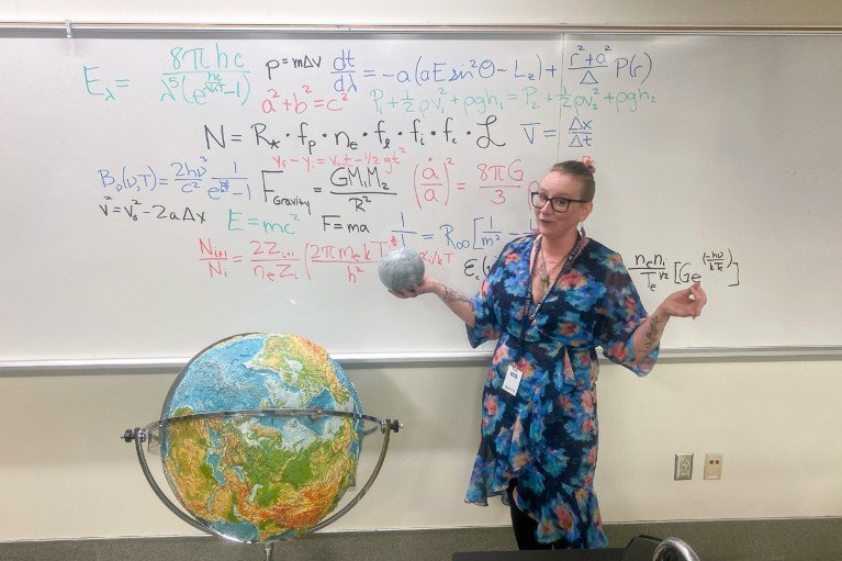 Profesora Seven Rasmussen enseña frente a una pizarra cubierta de ecuaciones, mientras está junto a un modelo de la Tierra y sostiene un modelo de la Luna
