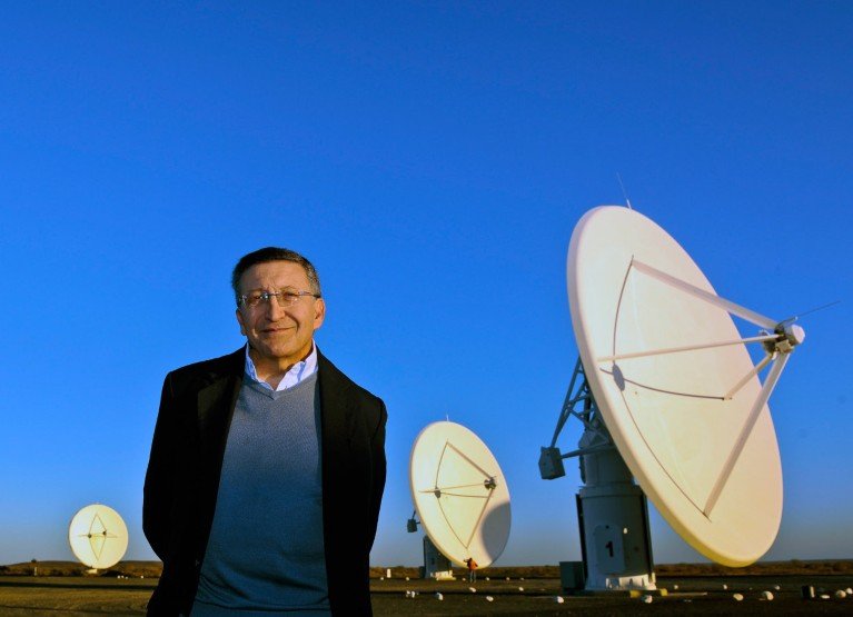 El Dr. Bernie Fanaroff sonriendo y de pie frente a un conjunto de antenas satelitales en Sudáfrica, con un cielo azul arriba de él.