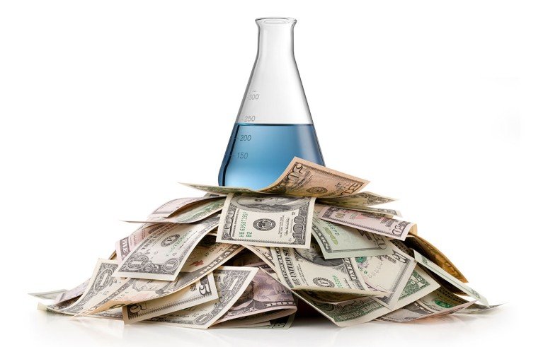 Un matraz Erlenmeyer con líquido azul sobre un montón de billetes de diferentes denominaciones de los Estados Unidos.